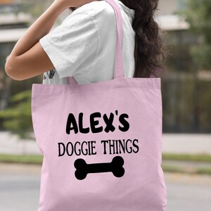 A woman wearing a white shirt and carrying a pink tote bag with the text "ALEX'S DOGGIE THINGS" printed on it.