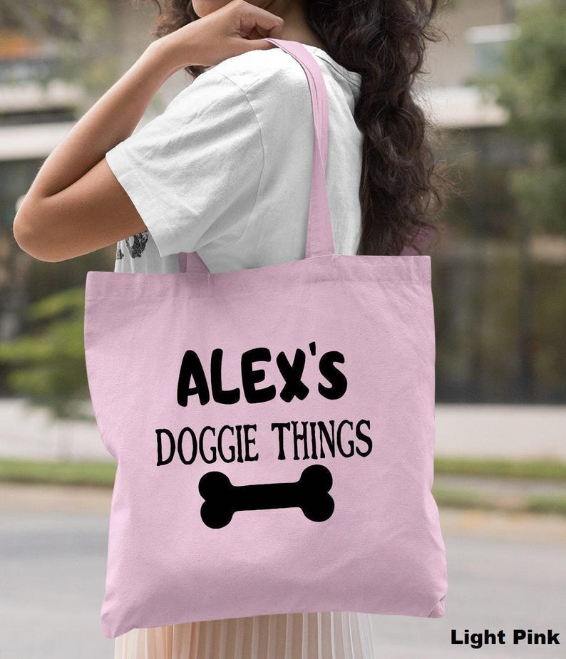 A woman wearing a white shirt and carrying a pink tote bag with the text "ALEX'S DOGGIE THINGS" printed on it.