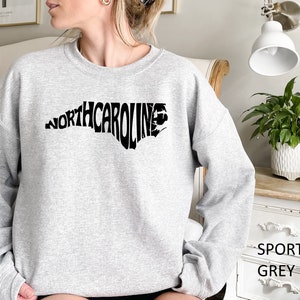 K&ouml;nnte beinhalten: Ein graues Sweatshirt mit einer schwarzen Grafik des Bundesstaates North Carolina, wobei das Wort "NORTHCAROLINE" in Form des Bundesstaates ausgeschrieben ist. Das Sweatshirt ist mit "SPORT GREY" gekennzeichnet.