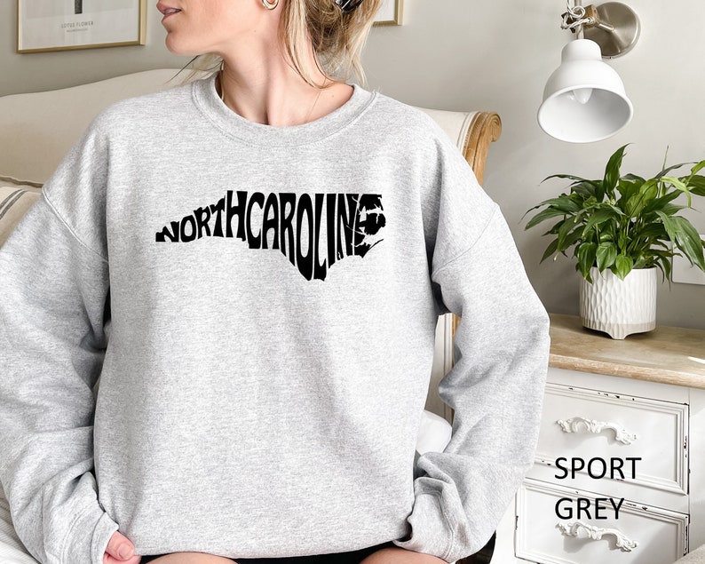 K&ouml;nnte beinhalten: Ein graues Sweatshirt mit einer schwarzen Grafik des Bundesstaates North Carolina, wobei das Wort "NORTHCAROLINE" in Form des Bundesstaates ausgeschrieben ist. Das Sweatshirt ist mit "SPORT GREY" gekennzeichnet.