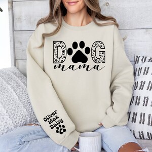 May include: A cream-colored sweatshirt with the text "DOG mama" in black lettering. The "D" and "G" are decorated with paw prints and bones. The sweatshirt has a black paw print on the left sleeve with the names "Oliver, Max, Daisy" written above it.