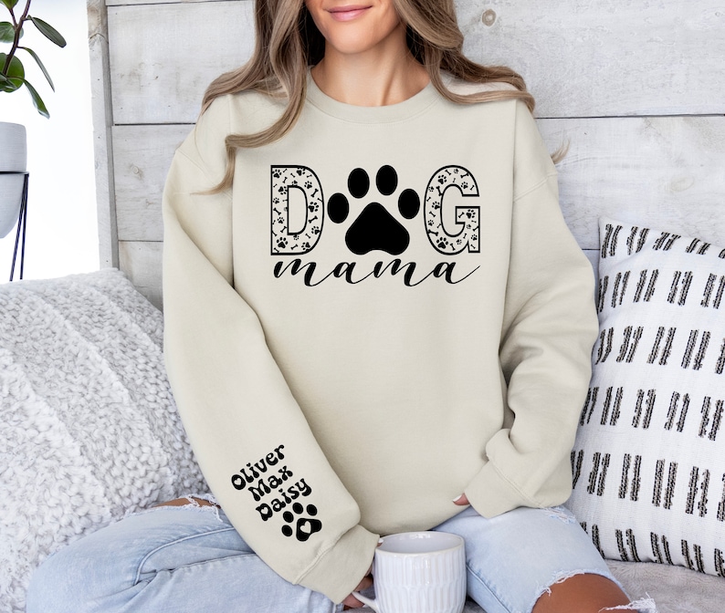 May include: A cream-colored sweatshirt with the text "DOG mama" in black lettering. The "D" and "G" are decorated with paw prints and bones. The sweatshirt has a black paw print on the left sleeve with the names "Oliver, Max, Daisy" written above it.