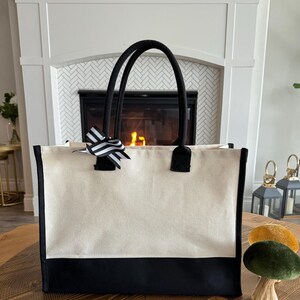 May include: A large, white canvas tote bag with black trim and black handles. The bag has a black bottom and a black and white striped bow on the front.