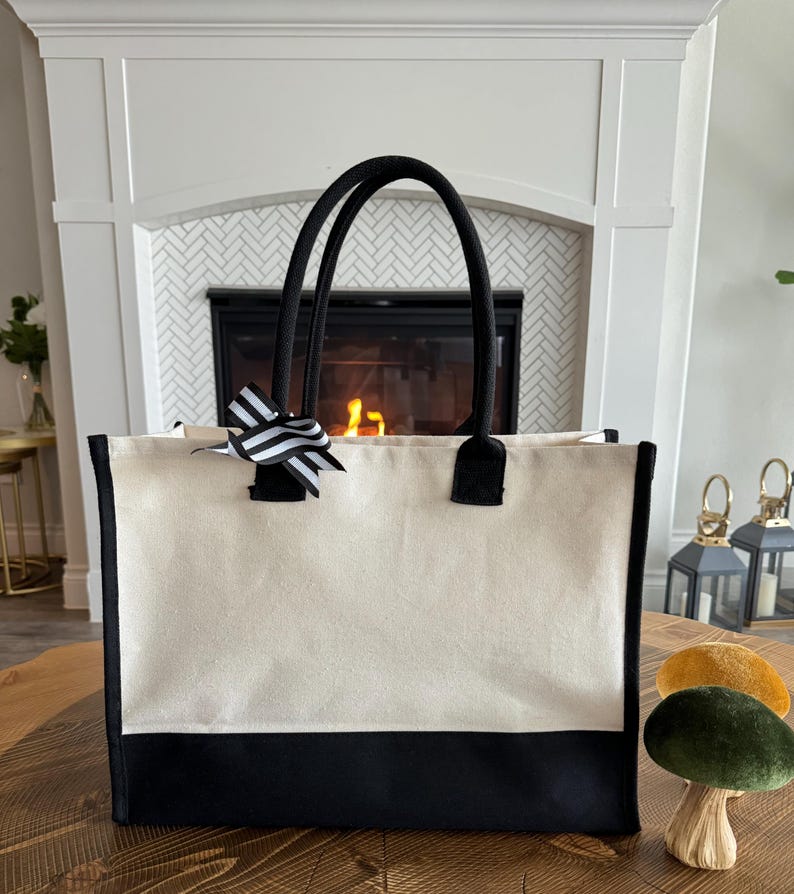 May include: A large, white canvas tote bag with black trim and black handles. The bag has a black bottom and a black and white striped bow on the front.