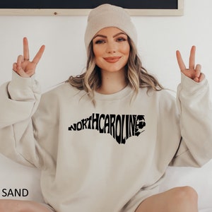 K&ouml;nnte beinhalten: Ein sandfarbener Pullover mit einem schwarzen Aufdruck des Bundesstaates North Carolina, wobei das Wort "NORTH CAROLINA" in Form des Bundesstaates ausgeschrieben ist.