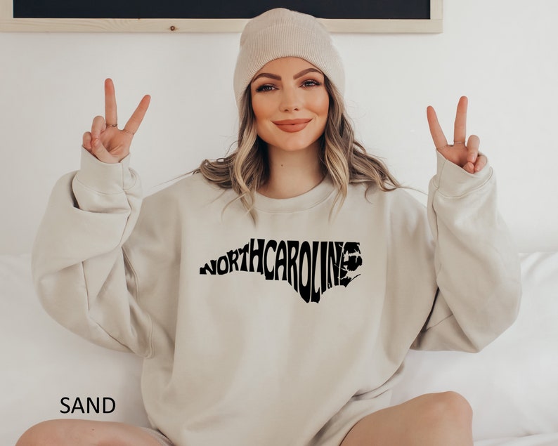 K&ouml;nnte beinhalten: Ein sandfarbener Pullover mit einem schwarzen Aufdruck des Bundesstaates North Carolina, wobei das Wort "NORTH CAROLINA" in Form des Bundesstaates ausgeschrieben ist.