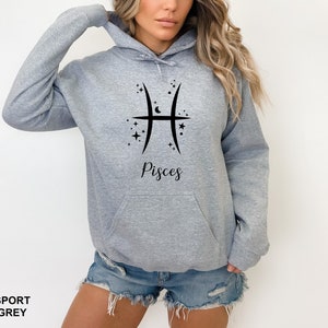 Op de afbeelding: Een grijs hoodie met een zwart Vissen sterrenbeeld en het woord "Vissen" gedrukt op de voorkant.