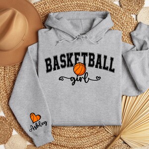May include: Gray hooded sweatshirt with the text "Basketball girl" and a basketball graphic. The sleeve has a heart graphic and the text "Ashley".