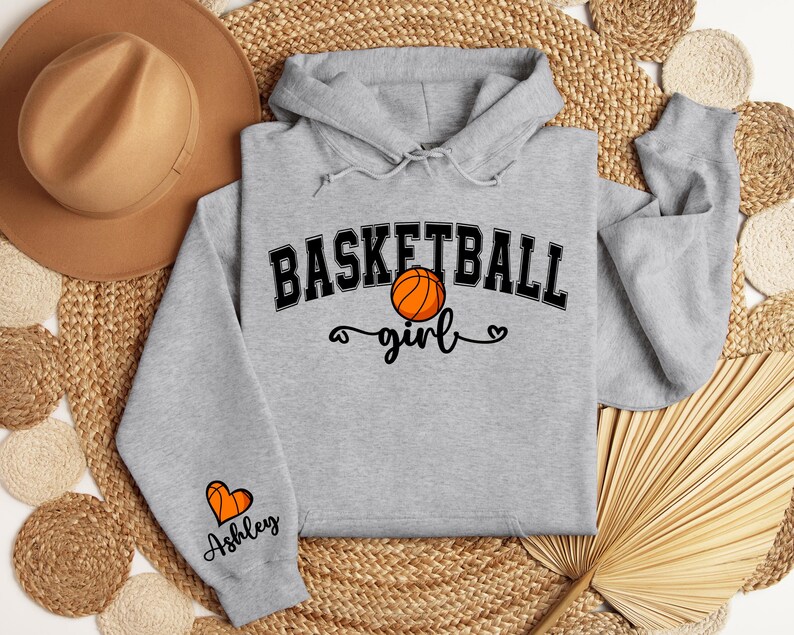 May include: Gray hooded sweatshirt with the text "Basketball girl" and a basketball graphic. The sleeve has a heart graphic and the text "Ashley".