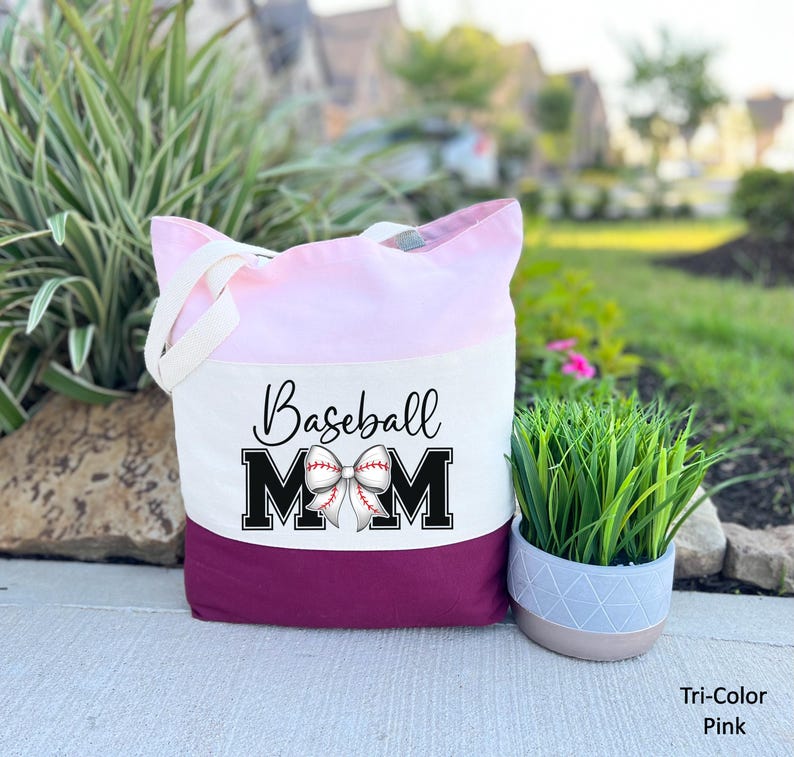May include: A pink and burgundy tote bag with the text "Baseball Mom" in black letters and a white baseball with a red bow on top.