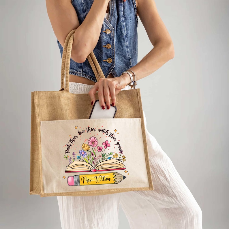 May include: A beige tote bag with a pocket on the front. The pocket has a design of an open book with flowers and a pencil. The text on the bag reads "Teach them, love them, watch them grow. Mrs. Wilson."