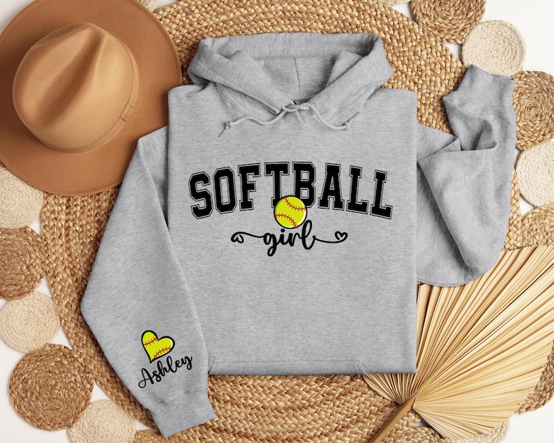 May include: Gray softball hoodie with the text "Softball girl" and a softball graphic. The left sleeve has a heart with the name "Ashley" on it.
