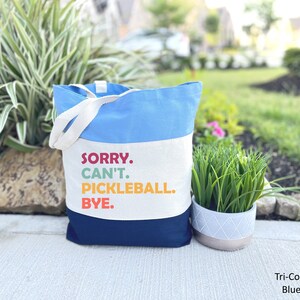 May include: A tri-color tote bag with a light blue top, white center, and navy blue bottom. The bag features white handles and the text "SORRY. CAN'T. PICKLEBALL. BYE." in colorful letters. A fun accessory for pickleball enthusiasts.