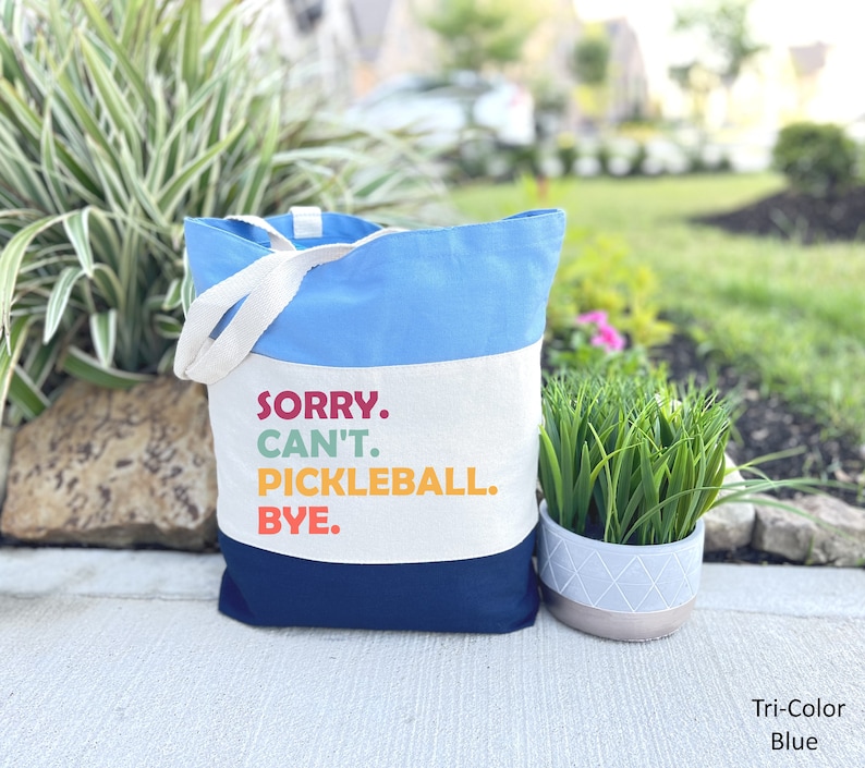 May include: A tri-color tote bag with a light blue top, white center, and navy blue bottom. The bag features white handles and the text "SORRY. CAN'T. PICKLEBALL. BYE." in colorful letters. A fun accessory for pickleball enthusiasts.