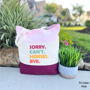 K&ouml;nnte beinhalten: Eine rosa und burgunderfarbene Canvas-Tragetasche mit dem Text "SORRY. CAN'T. HORSES. BYE." in bunten Buchstaben. Die Tasche hat lange Henkel und steht auf einem B&uuml;rgersteig.
