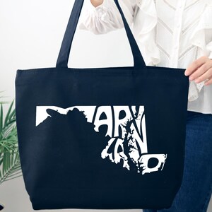 May include: A navy blue tote bag with white text and a graphic of the state of Maryland. The bag has two long handles and is held by a person. The text reads "MARYLAND".