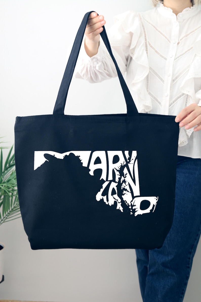 May include: A navy blue tote bag with white text and a graphic of the state of Maryland. The bag has two long handles and is held by a person. The text reads "MARYLAND".