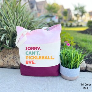 May include: A tri-color pink tote bag with the text "SORRY. CAN'T. PICKLEBALL. BYE." in colorful letters. The bag has a pink top, off-white middle section, and a deep magenta bottom. It has off-white handles.