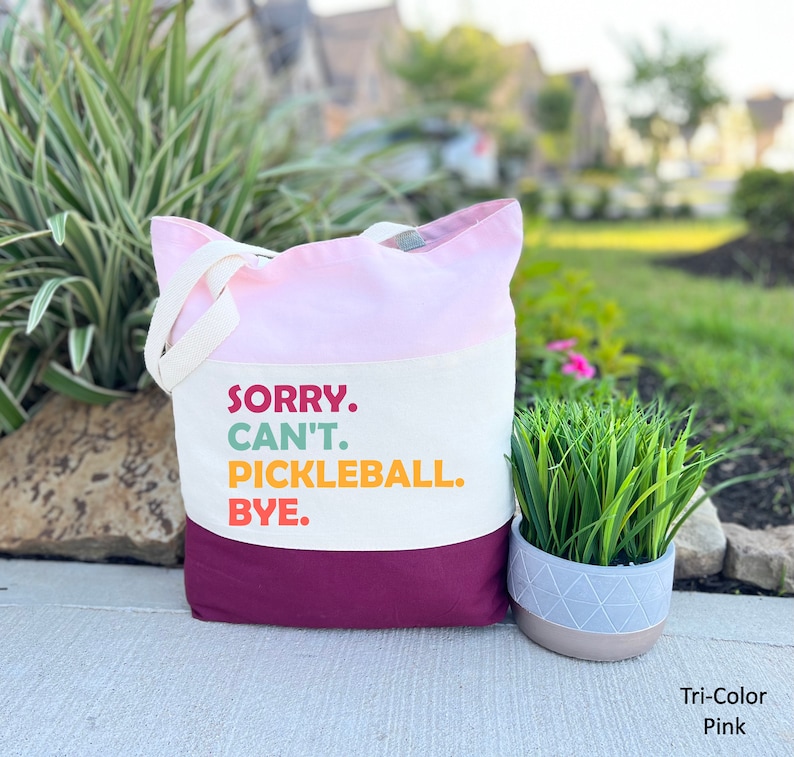 May include: A tri-color pink tote bag with the text "SORRY. CAN'T. PICKLEBALL. BYE." in colorful letters. The bag has a pink top, off-white middle section, and a deep magenta bottom. It has off-white handles.