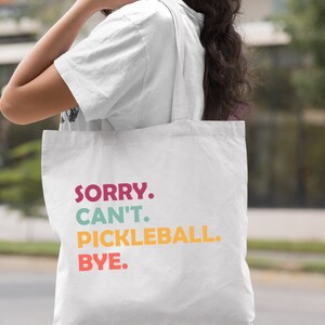 May include: White tote bag with the text "SORRY. CAN'T. PICKLEBALL. BYE." in colorful letters. The bag is being carried over the shoulder.