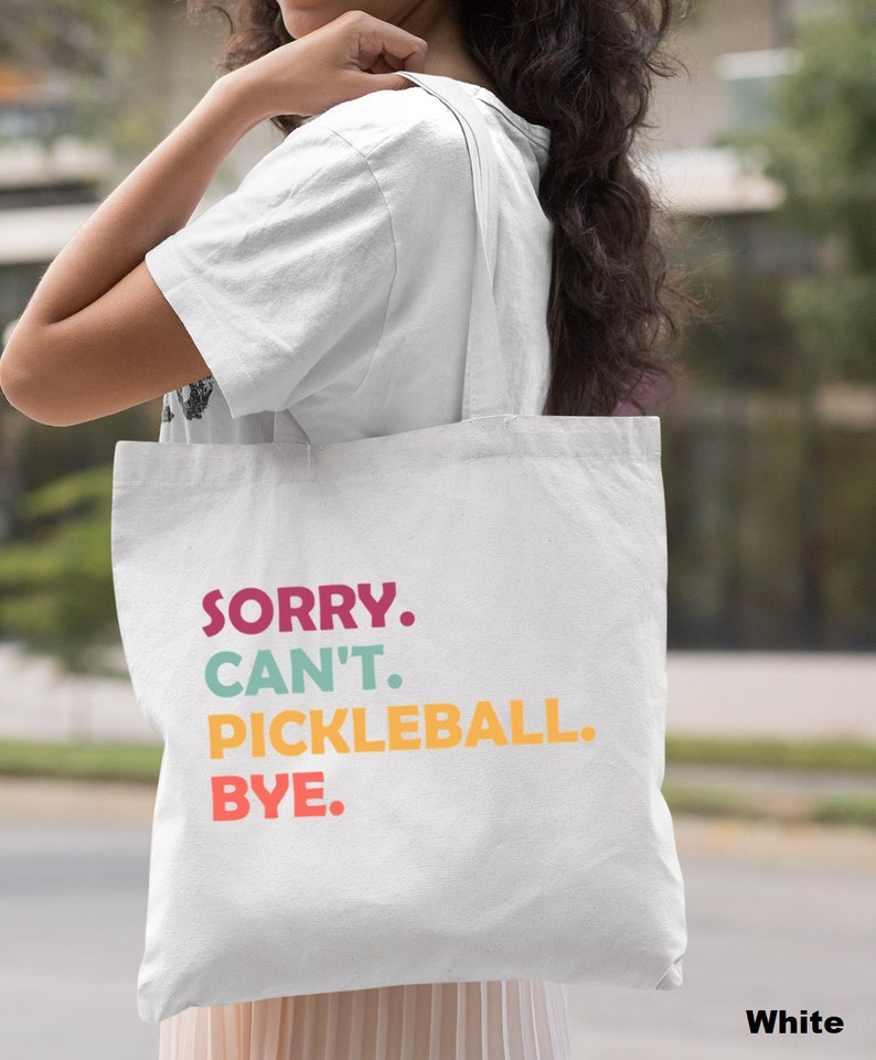 May include: White tote bag with the text "SORRY. CAN'T. PICKLEBALL. BYE." in colorful letters. The bag is being carried over the shoulder.