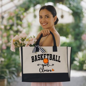 May include: A canvas tote bag with black handles and a black bottom panel. The bag features the text "BASKETBALL girl Claire" with a basketball graphic and heart accents. A black and white striped bow is tied to the handle.