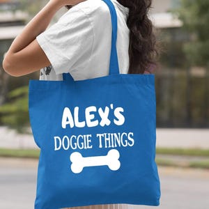 A woman is holding a blue tote bag with the text "ALEX'S DOGGIE THINGS" printed on it.