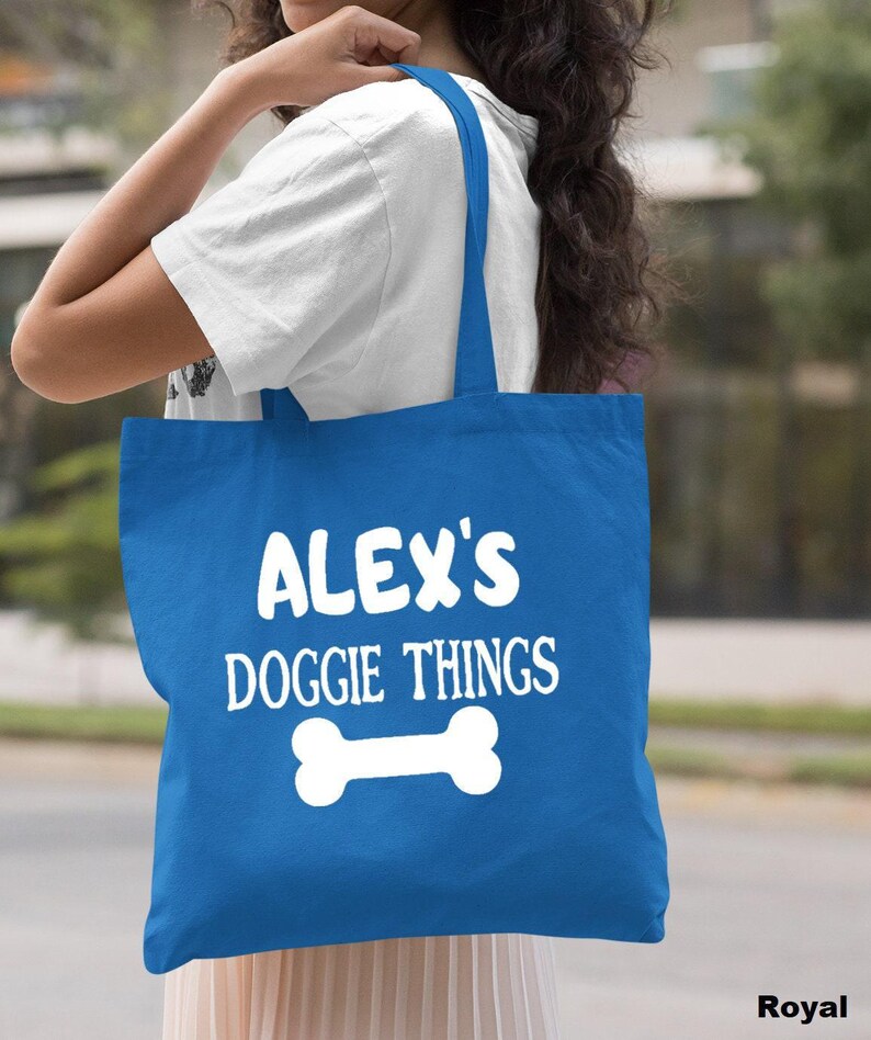 A woman is holding a blue tote bag with the text "ALEX'S DOGGIE THINGS" printed on it.