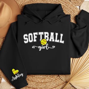 May include: Black hoodie with the text "Softball girl" in white with a yellow softball graphic. The left sleeve has a small heart with the name "Ashley" in white.
