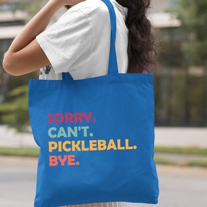 May include: Royal blue tote bag with the text "SORRY. CAN'T. PICKLEBALL. BYE." in red, teal, yellow, and orange. The bag has long straps and is made of a sturdy fabric. The bag is being held by a person.