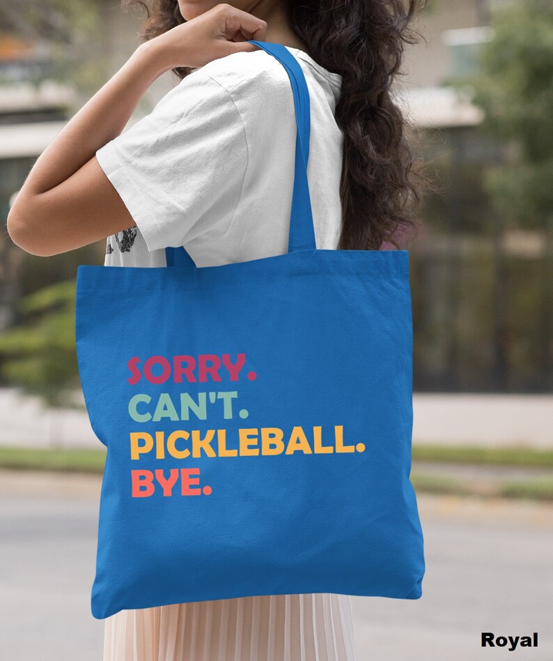 May include: Royal blue tote bag with the text "SORRY. CAN'T. PICKLEBALL. BYE." in red, teal, yellow, and orange. The bag has long straps and is made of a sturdy fabric. The bag is being held by a person.