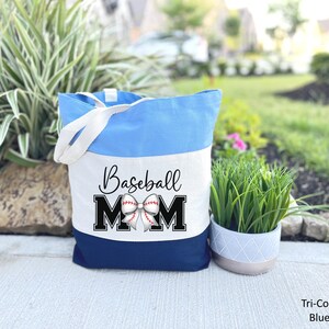 May include: A blue and white canvas tote bag with the text "Baseball Mom" and a baseball with a bow on it. The bag has a white strap.