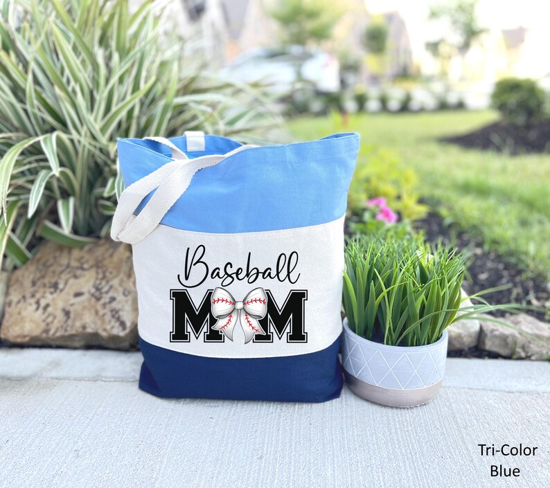 May include: A blue and white canvas tote bag with the text "Baseball Mom" and a baseball with a bow on it. The bag has a white strap.
