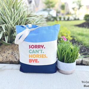 K&ouml;nnte beinhalten: Eine blaue, wei&szlig;e und marineblaue Canvas-Tragetasche mit dem Text "SORRY. CAN'T. HORSES. BYE." in bunten Buchstaben. Die Tasche hat zwei wei&szlig;e Canvas-Henkel.