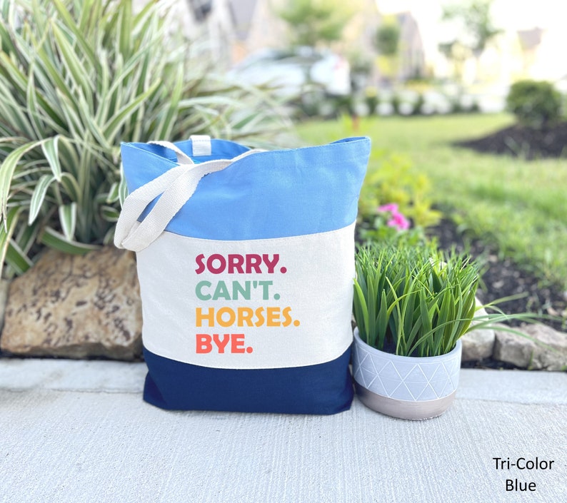 K&ouml;nnte beinhalten: Eine blaue, wei&szlig;e und marineblaue Canvas-Tragetasche mit dem Text "SORRY. CAN'T. HORSES. BYE." in bunten Buchstaben. Die Tasche hat zwei wei&szlig;e Canvas-Henkel.