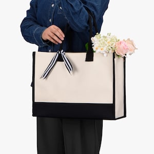 Puede incluir: Una persona sostiene una gran bolsa de lona blanca con ribetes negros. La bolsa tiene una cinta rayada en blanco y negro atada a las asas. La bolsa est&aacute; llena de un ramo de flores blancas y rosas.