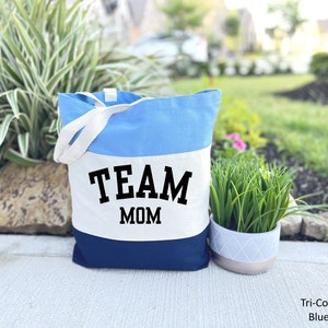 May include: A blue, white, and navy blue canvas tote bag with the text "TEAM MOM" in black.