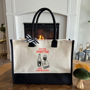 May include: Cream-colored tote bag with black accents and handles. The bag features the text "ONE MORE CHAPTER and MAYBE A GLASS." A black and white striped bow is attached to the handle. The bag has a black bottom.