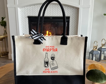 May include: Cream-colored tote bag with black accents and handles. The bag features the text "ONE MORE CHAPTER and MAYBE A GLASS." A black and white striped bow is attached to the handle. The bag has a black bottom.