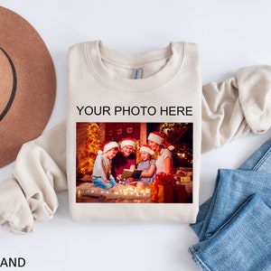 May include: A beige sweatshirt with the text "YOUR PHOTO HERE" printed on the front. A photo of a family of four wearing Santa hats is displayed in the space provided for a personalized photo. The photo shows the family sitting in front of a Christmas tree with lights.