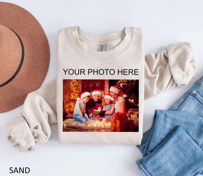 May include: A beige sweatshirt with the text "YOUR PHOTO HERE" printed on the front. A photo of a family of four wearing Santa hats is displayed in the space provided for a personalized photo. The photo shows the family sitting in front of a Christmas tree with lights.