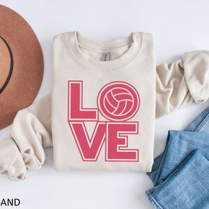 May include: A beige sweatshirt with the word "LOVE" in pink letters with a volleyball graphic in the "O".