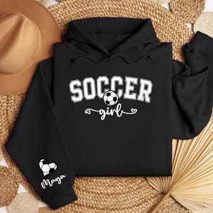 May include: Black hoodie with a white graphic that says "Soccer girl" with a soccer ball and a heart. The left sleeve has a white heart with a soccer ball and the name "Maya" written below.