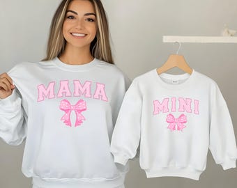 May include: White sweatshirts with pink text and bow designs. One sweatshirt says "MAMA" and the other says "MINI". The bows have a polka dot pattern. The sweatshirts are a matching set.