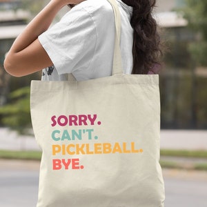 May include: A natural canvas tote bag with the text "SORRY. CAN'T. PICKLEBALL. BYE." in colorful letters. The bag has long straps and is held by a person. The word "Natural" is printed in the bottom right corner.