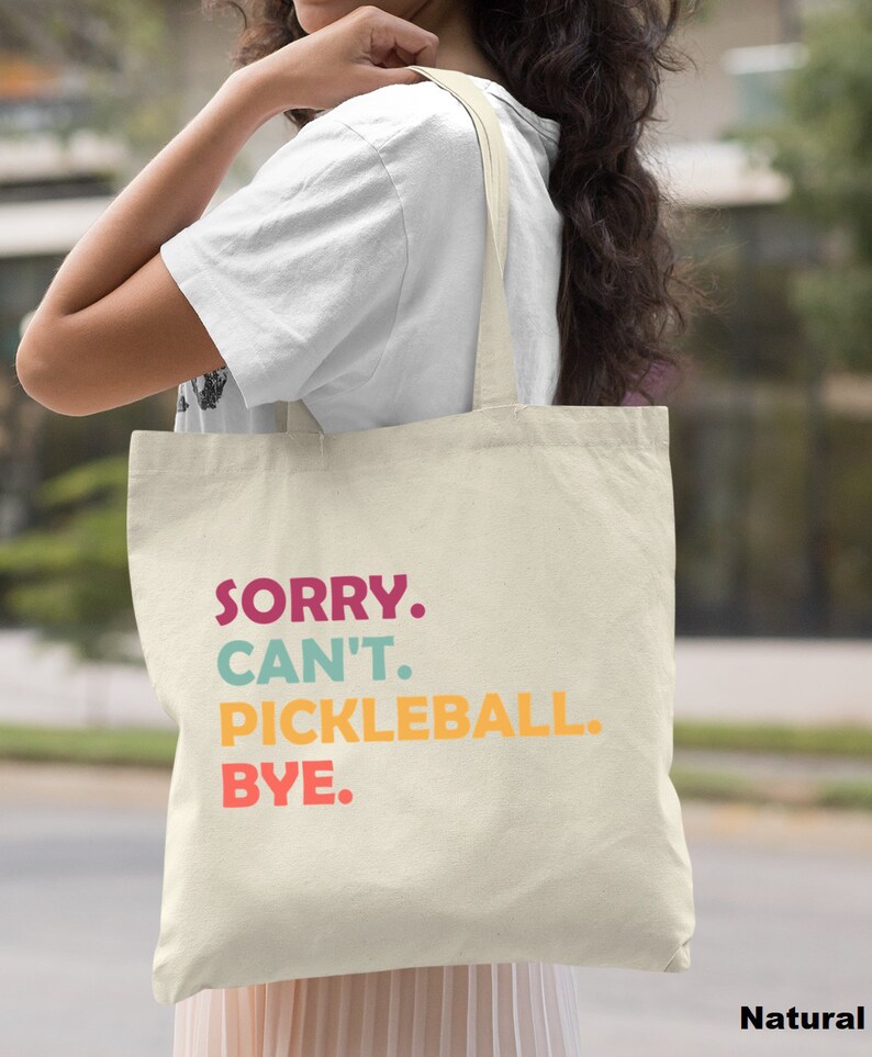 May include: A natural canvas tote bag with the text "SORRY. CAN'T. PICKLEBALL. BYE." in colorful letters. The bag has long straps and is held by a person. The word "Natural" is printed in the bottom right corner.