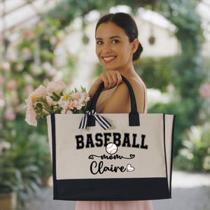 May include: A beige tote bag with black accents and handles. The bag features the text "BASEBALL mom Claire" with a baseball graphic and heart accents. A black and white striped bow is tied to the handle.