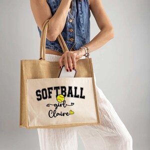 May include: A tan jute tote bag with a cream-colored front pocket. The pocket features the word "SOFTBALL" in black block letters, a yellow softball graphic, and the words "girl Claire" in script. The bag has tan handles.