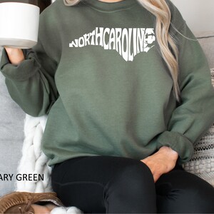 K&ouml;nnte beinhalten: Ein olivgr&uuml;nes Sweatshirt mit einer wei&szlig;en Umrandung des Bundesstaates North Carolina, wobei das Wort "NORTH CAROLINA" innerhalb der Umrandung ausgeschrieben ist.