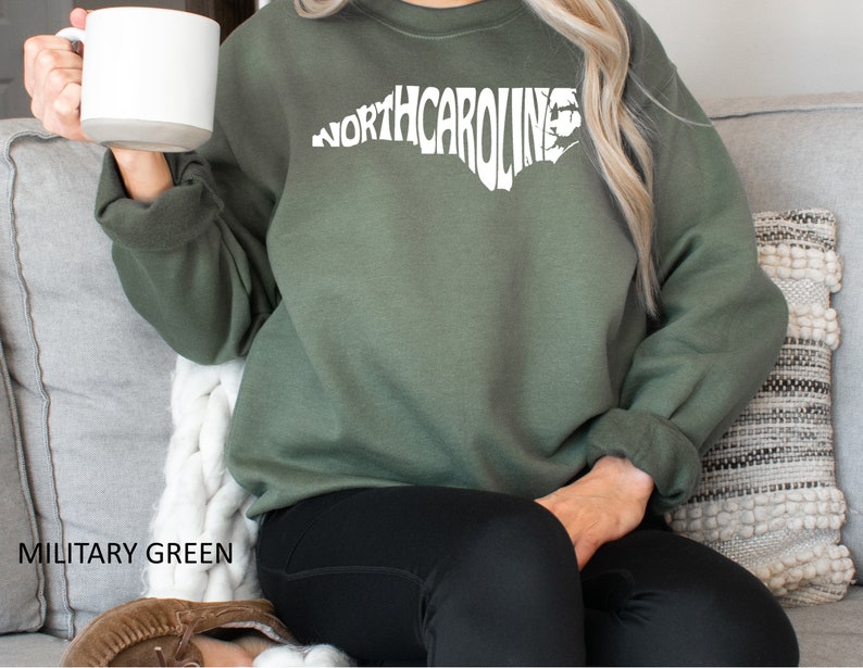 K&ouml;nnte beinhalten: Ein olivgr&uuml;nes Sweatshirt mit einer wei&szlig;en Umrandung des Bundesstaates North Carolina, wobei das Wort "NORTH CAROLINA" innerhalb der Umrandung ausgeschrieben ist.